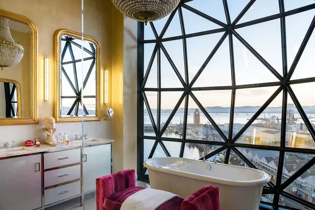 Imagem de banheira, pia com dois espelhos, puff e janela do Hotel Silo na Cidade do Cabo. 